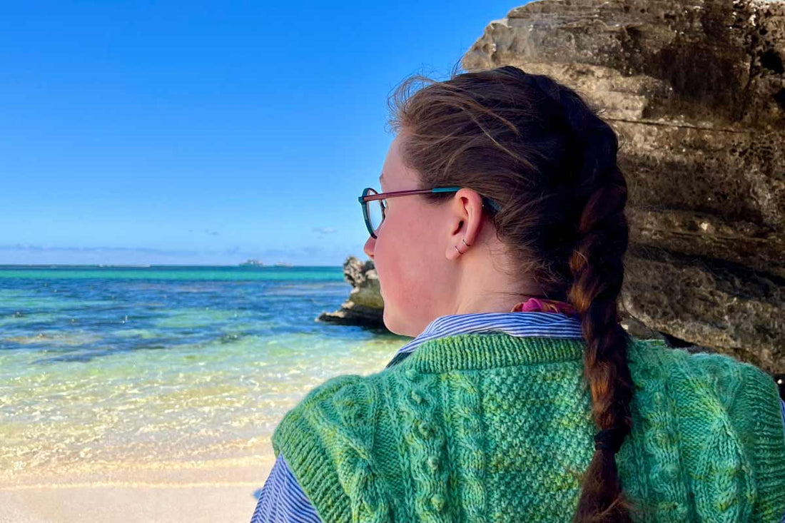 Alex, a white woman in her late 20s, looks out to sea while sat on a sandy beach on a clear and sunny day in Australia.