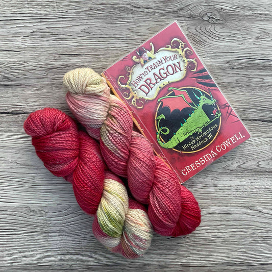 Two skeins of red yarn beside a book of How To Train Your Dragon, the cover is the same colours as the yarn.