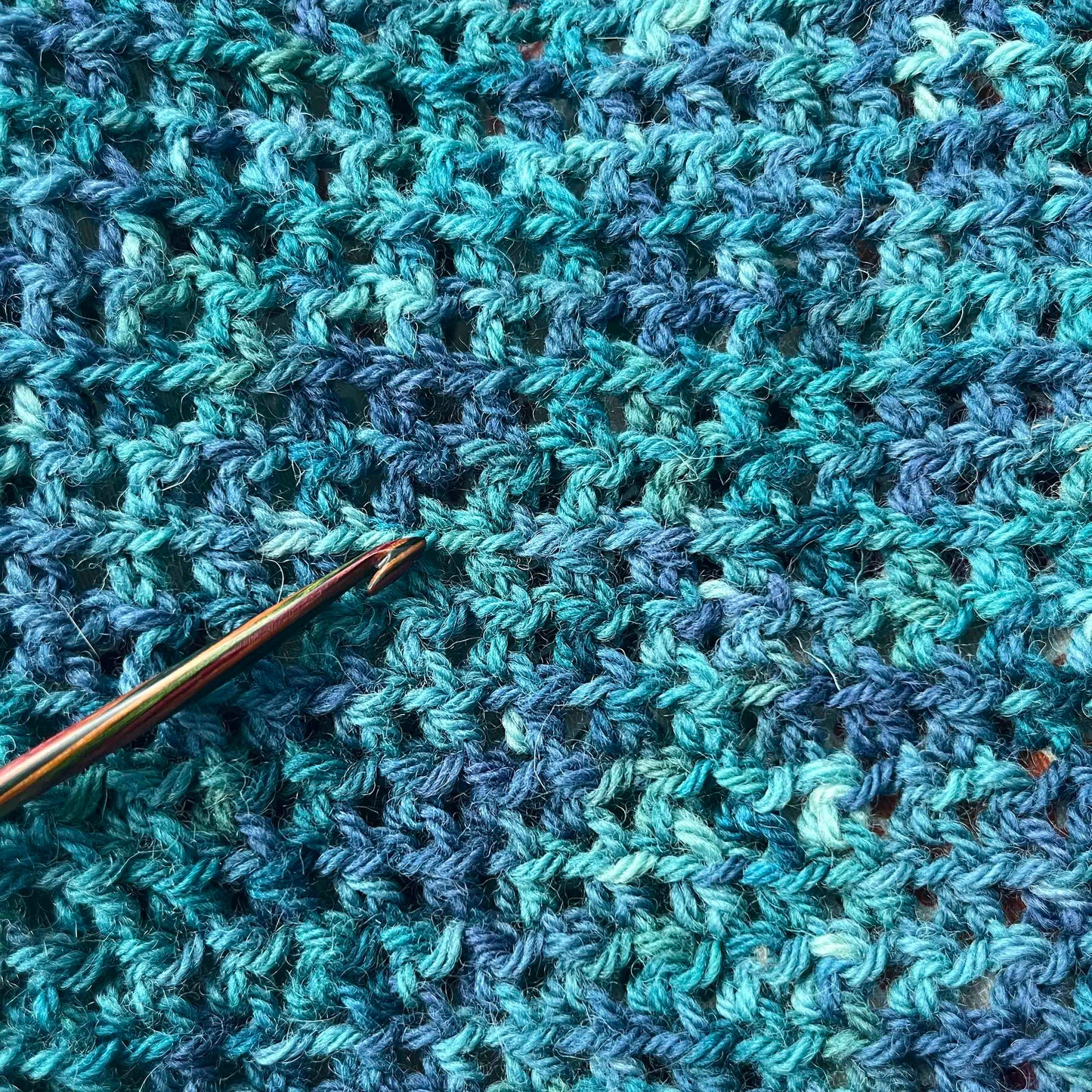 A close up of crochet, with a crochet hook resting on the work. The yarn is dark teals, blues and greens.