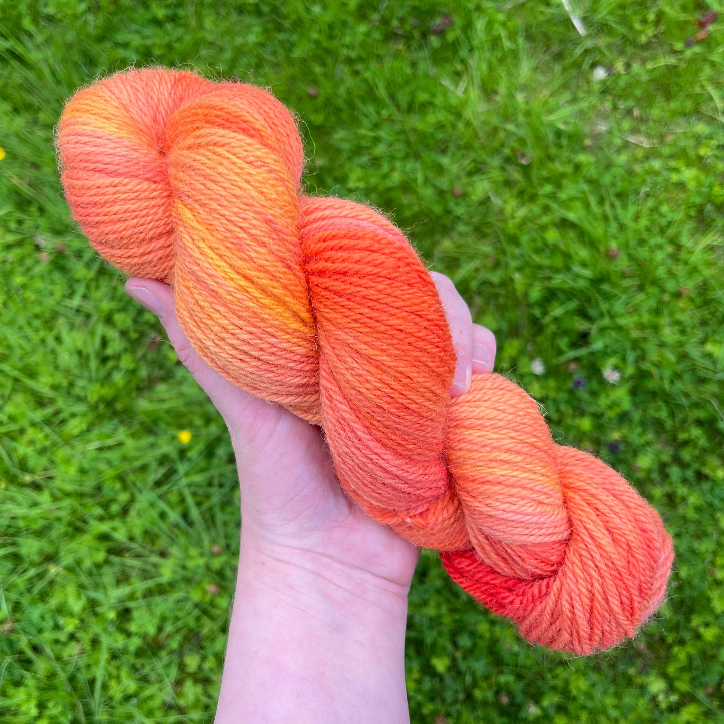 A white hand holds a skein of tonal peach yarn over fresh green grass.