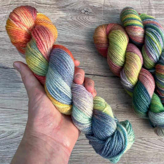 A white hand holds a skein of muted rainbow yarn.