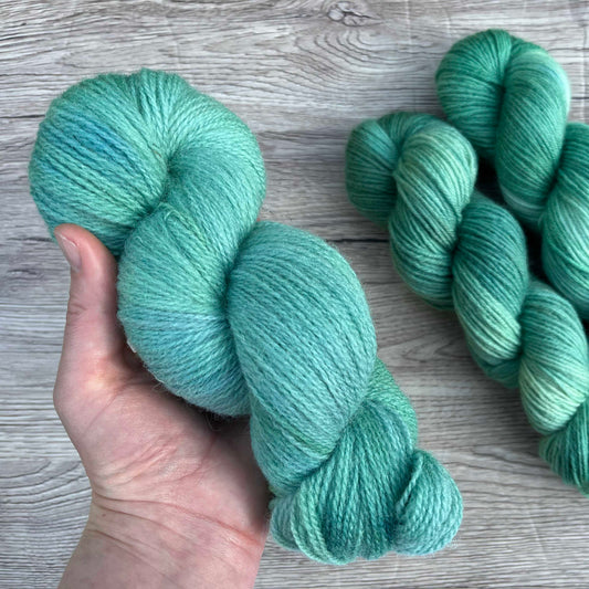 A white hand holds a skein of teal yarn to the camera. 