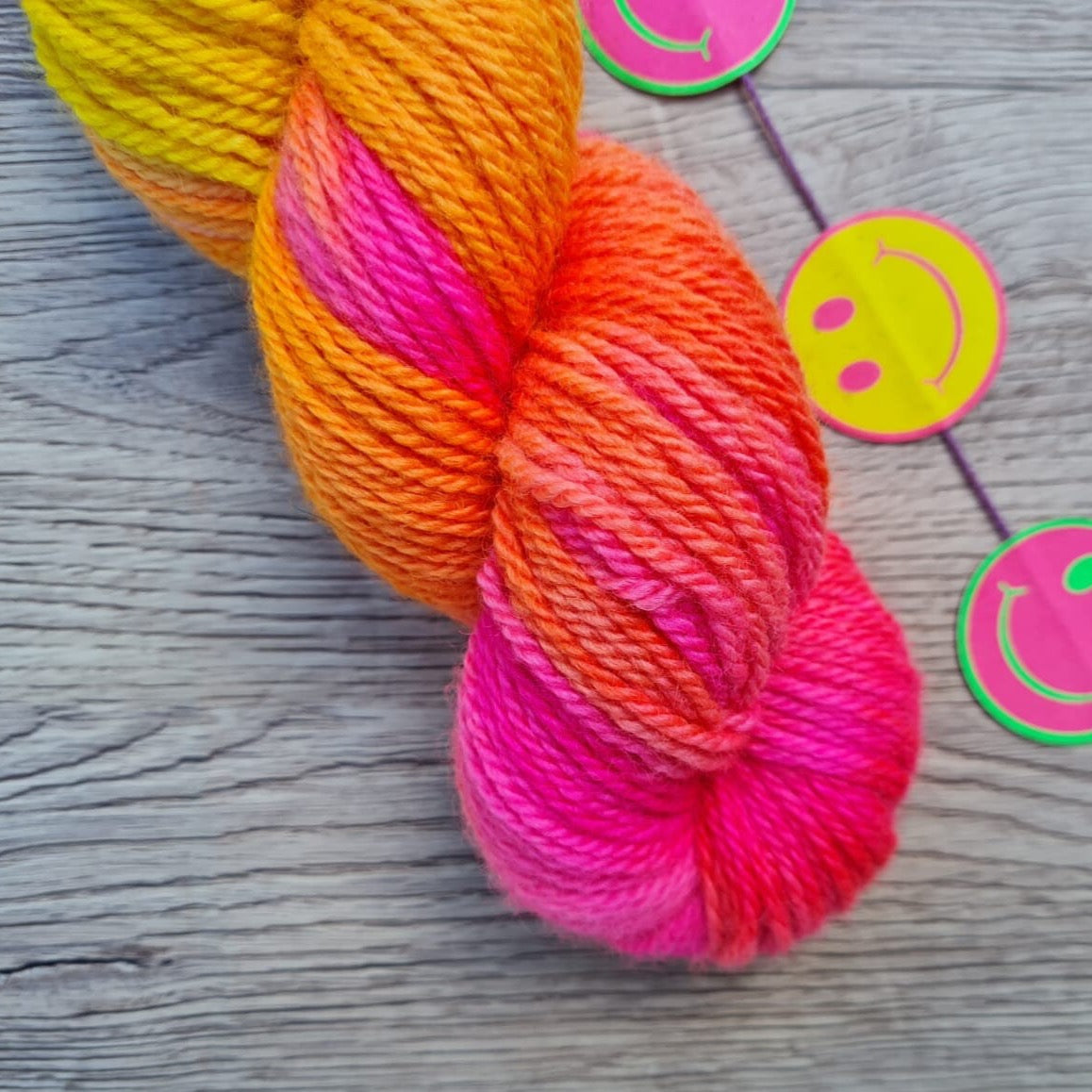 A close up of a skein of bright pink and yellow yarn next to smiley face stickers in the same colours.