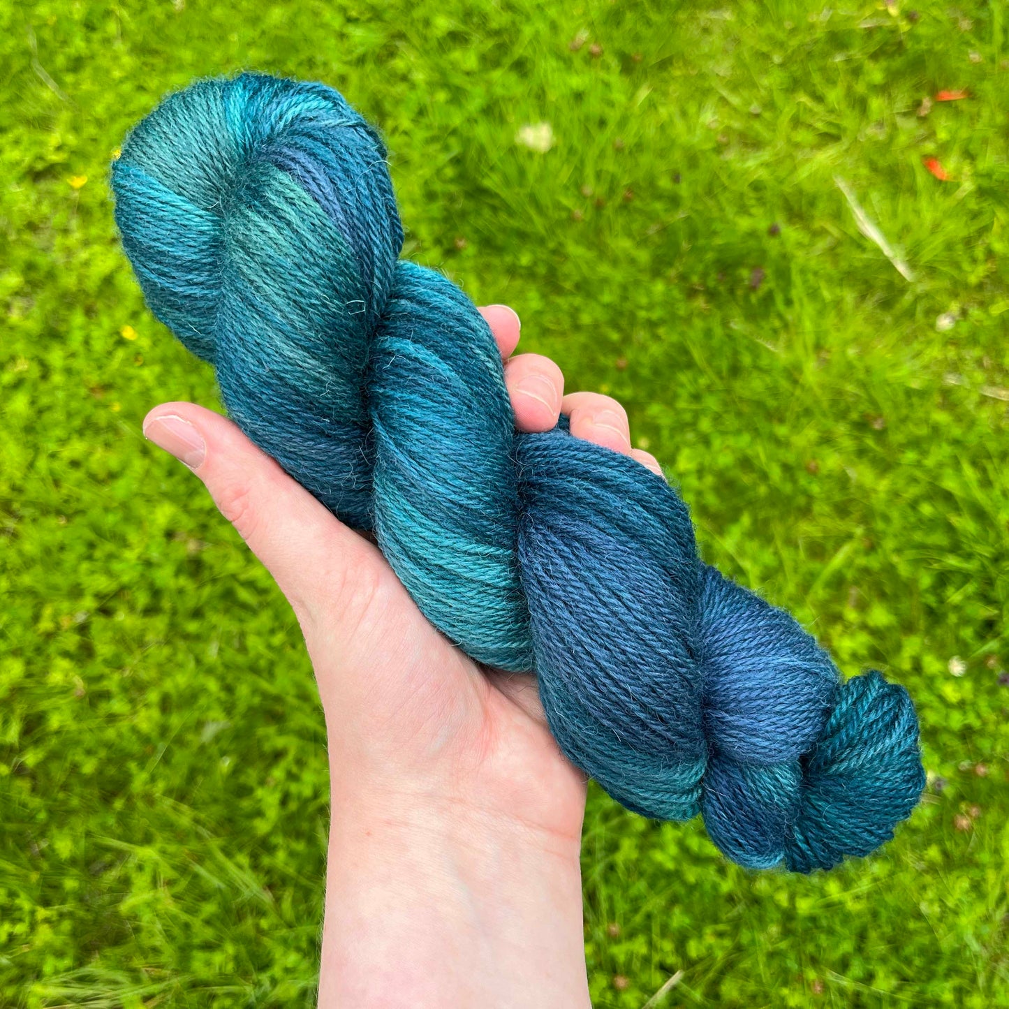 A white hand holds a skein of dark teal, green and blue yarn over fresh green grass.