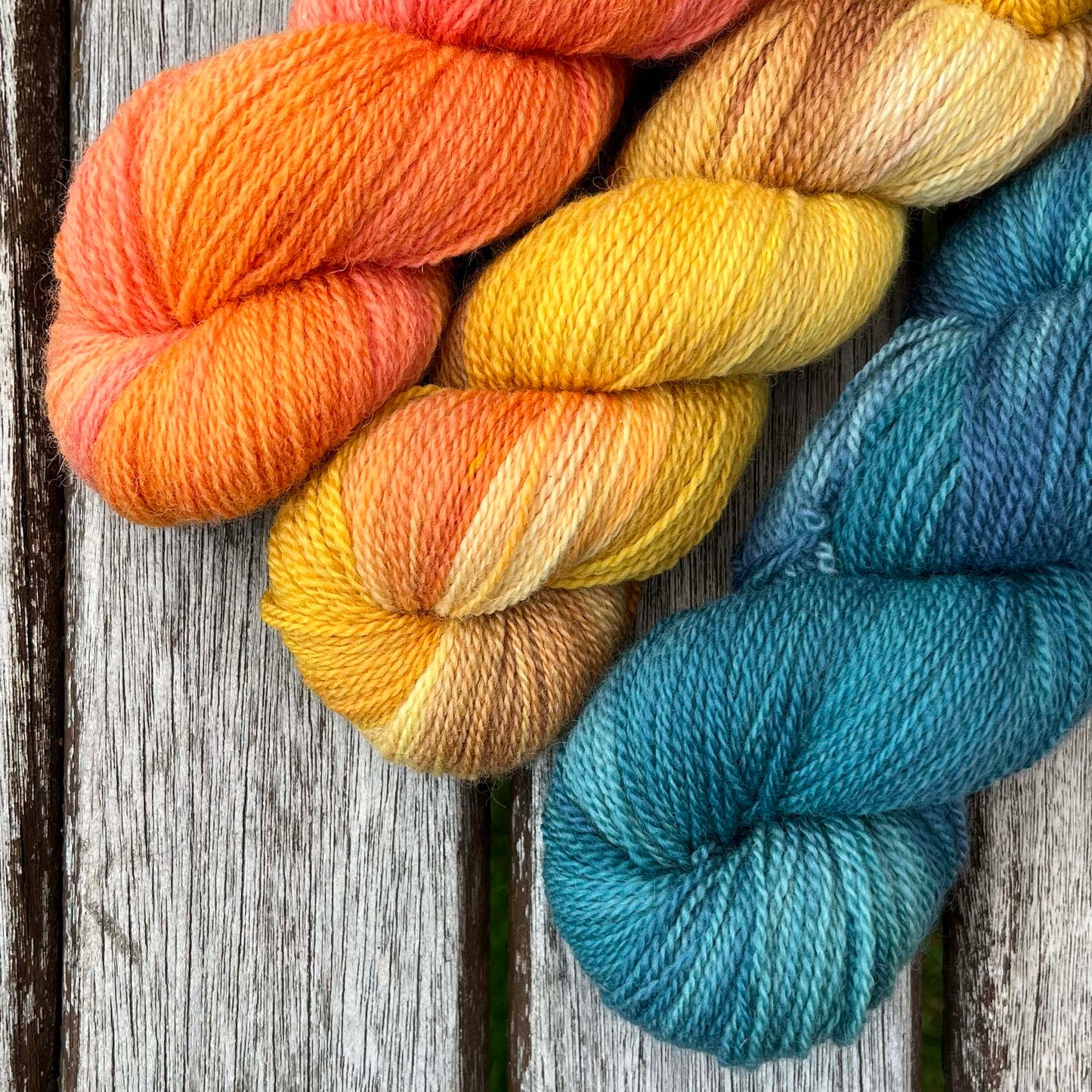 Three skeins of yarn peek over a wood background, one peach, one golden, one teal.
